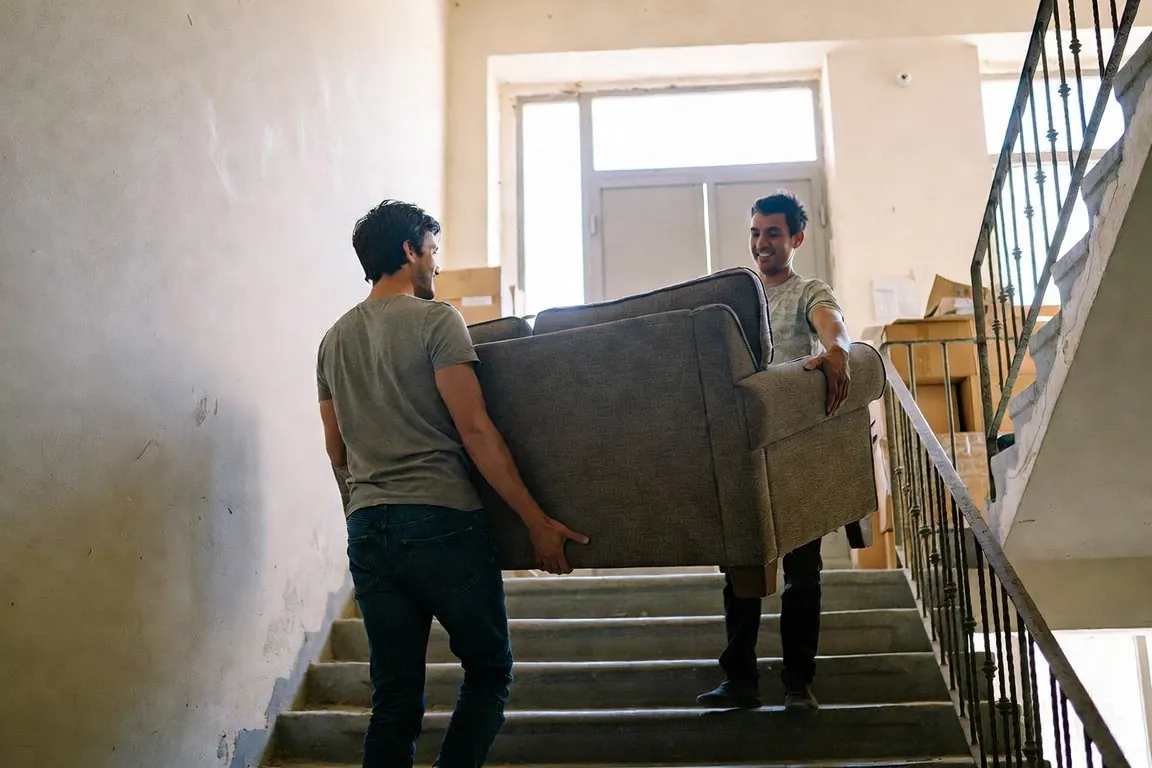 Two friends carry a sofa up an apartment stairwell past stacked moving boxes